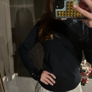 Svart långärmad tröja med knappar - Snygg svart långärmad tröja med dekorativa knappar vid ärmsluten. Tröjan har en normal passform och ett mjukt material. Från Lindex. Tröjan är mörkblå.