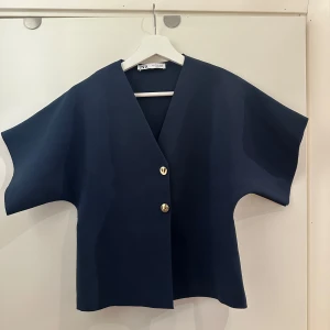 Tröja - Snygg mörkblå jacka med korta ärmar och två dekorativa knappar framtill. Jackan har en v-ringad design och en stilren look. Perfekt för en trendig och avslappnad stil.