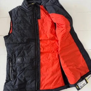 Svart smart uppvärmd väst från Blåkläder - Helt ny i kartong! Mörkblå smart uppvärmd väst från Blåkläder med dragkedja och röd insida. Perfekt för kyliga dagar med sin innovativa uppvärmningsteknik. Västen har en praktisk ficka med dragkedja och kommer med laddare och batteri.Väst som med sin smarta värmeteknik från Clim8® känner av när din kroppstemperatur sjunker och ser till att värma upp dig. Quiltad på både bål och rygg samt har stickad polyester i sidorna, vilket bidrar till västens extraordinära rörlighet och komfort. 3760 nypris.