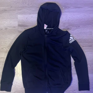 Svart huvtröja från Adidas - Säljer en svart huvtröja från Adidas med dragkedja och logga på ärmen. Tröjan har långa ärmar och en bekväm huva. Perfekt för sportiga dagar eller avslappnade stunder.