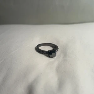 Svart ring med sten - Elegant simpel svart ring med en stor, mörk sten i mitten. Ringen har en detaljerad design med små stenar längs bandet som ger extra glans och stil. Perfekt för att ge en sofistikerad touch till din look.