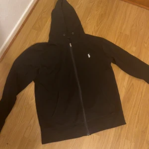 Svart huvtröja från Ralph Lauren - Säljer en stilren svart huvtröja från Ralph Lauren med dragkedja och det klassiska logotypbroderiet på bröstet. Tröjan har långa ärmar och en praktisk huva. Perfekt för en avslappnad look.