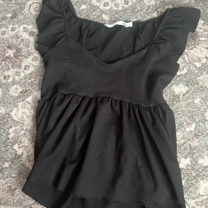 Svart topp från 157 - Snygg svart topp från 157 med volangdetaljer vid axlarna. Toppen har en v-ringad design och är tillverkad i ett ribbat material som ger en fin struktur. Har kanska använt 2 gånger 