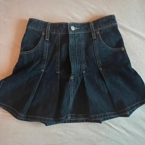 Mörkblå jeanskjol - Trendig mörkblå jeanskjol med veckade detaljer och klassiska fickor fram. Kjolen har en knapp och dragkedja i midjan för enkel påklädning. Perfekt för en avslappnad stil.