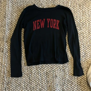 Svart långärmad tröja med New York-tryck - Snygg svart tröja med rött 'New York'-tryck på framsidan. Tröjan har långa ärmar och en bekväm passform, perfekt för en avslappnad stil.