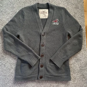 Stickad cardigan - Säljer en stilren grå kofta från Hollister med broderad logga och årtal 1922 på bröstet. Koftan har en v-ringad design med knappar och ribbade ärmslut. Perfekt för en avslappnad look. Skick 10/10 in princip ny. Modellen är 186. Skriv för fler frågor eller bilder. Perfekt nu till våren!