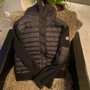 Svart pufferjacka från Moncler - Säljer en snygg svart pufferjacka från Moncler. Jackan har en quiltad design med dragkedja framtill och långa ärmar. Den är perfekt för kyligare dagar och har en stilren look med ribbade detaljer. Storleken är L men den sitter mer som en M, passar även de med storlek L beroende på kroppstyp.