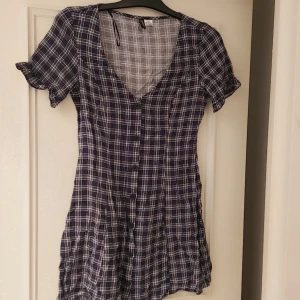 Rutig klänning från H&M - Säljer en snygg rutig klänning från H&M i mörkblå och vit. Klänningen har korta ärmar och en v-ringad design med knappar framtill. Perfekt för en avslappnad stil.