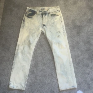 Acid wash jeans från Levi's - Säljer ett par coola acid wash jeans från Levi's, modell 501. De har en unik blekt look med klassisk femficksdesign och knappgylf. Perfekta för dig som vill sticka ut med en retro stil. Passar väl till vitt. Har även en ”bullet hole” effekt på ena benet där små hål går runt på yttersidan av benet. Sitter bra på mig som gillar 32x32~34. Står w34 men upplever 32~33. 
