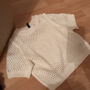 Vit stickad tröja - Säljer en vit stickad tröja med ett luftigt hålmönster. Tröjan har korta ärmar och ribbade kanter vid ärmslut och nederkant. Perfekt för lager-på-lager-stil eller sommarens svalare dagar.