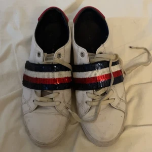 Vita sneakers från Tommy Hilfiger med paljettdetaljer - Säljer dessa skit Snygga vita sneakers från Tommy Hilfiger med en bred rem dekorerad med paljetter i rött, vitt och blått. Skorna har snörning och en röd detalj vid hälen. Säljer dom då de tyvärr har blivit för små för mig. Dom är i mycket bra skick men kan behöva torkas av. 