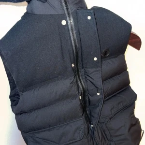 Svart dunväst från Moncler - Nu har vi fått in den igen!!! Säljer en stilren svart dunväst från Moncler med dragkedja och knappar framtill. Västen har en hög krage och Moncler-logga på bröstet. 