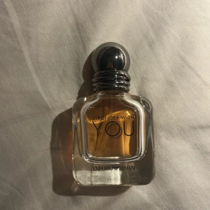 Stronger With You parfym från Emporio Armani - Säljer en elegant parfymflaska av märket Emporio Armani, 'Stronger With You'. Flaskan har en stilren design med en rundad silverfärgad kork och genomskinligt glas som visar den gyllene doften inuti. Perfekt för den som vill ha en sofistikerad och modern doft.