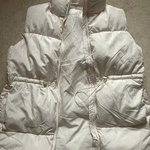 Vit dunväst från Amisu - Säljer en stilren vit dunväst från Amisu. Västen har en dragkedja framtill och praktiska fickor på sidorna. Perfekt för kyligare dagar när du vill hålla värmen utan att kompromissa med stilen.