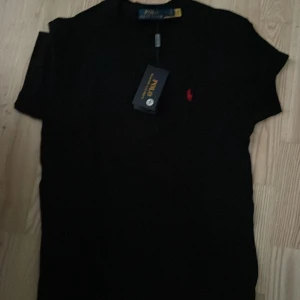 Svart t-shirt från Ralph Lauren - Snygg svart t-shirt från Ralph Lauren med det klassiska röda broderade logotypen på bröstet. Perfekt för en stilren look. T-shirten är kortärmad och gjord i mjukt material för extra komfort.