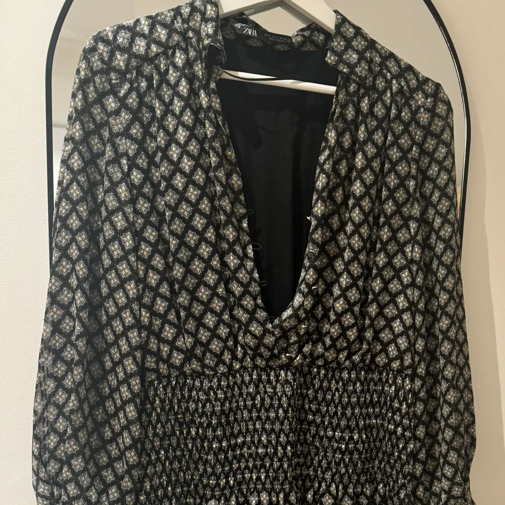 Snygg mönstrad klänning från Zara med långa ärmar och v-ringning. Klänningen har ett smockat midjeparti och ett geometriskt mönster i svart och vitt. Perfekt för en stilfull look.. Mekot.