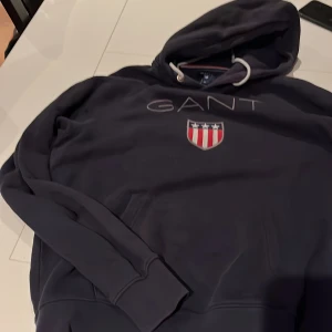 Mörkblå hoodie från GANT - Säljer en snygg mörkblå hoodie från GANT med deras ikoniska logga och ett broderat emblem på bröstet. Tröjan har långa ärmar och en klassisk huva med dragsko. Perfekt för en avslappnad stil.