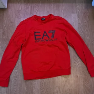 Röd tröja från Emporio Armani - Snygg röd tröja från Emporio Armani med EA7-logga på framsidan. Tröjan har långa ärmar och en klassisk rund halsringning. Perfekt för en stilren och avslappnad look. St ungefär om man är 160 eller 170cm lång 