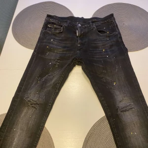 Svarta jeans med slitningar och färgstänk - Coola svarta jeans med slitningar och unika färgstänk. De har en klassisk femficksdesign och knappgylf. Perfekta för en avslappnad stil med en twist.