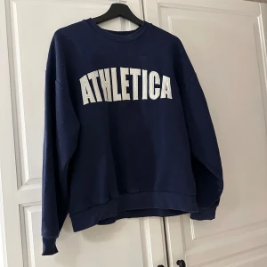 Mörkblå sweatshirt från Gina Tricot - Säljer en mörkblå sweatshirt från Gina Tricot med texten 'ATHLETICA' i vitt på framsidan. Tröjan har långa ärmar och en rund halsringning. Perfekt för en avslappnad stil. 🖤