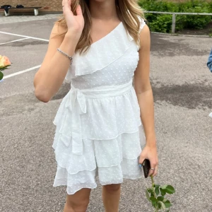 Vit studentklänning från Showpo - Säljer denna söta klänningen med volanger. Klänningen har en oneshoulder och knytband på axeln och i midjan. Strl 38, men passar mig som har strl 34/36 då man kan spänna bandet både i midjan och axeln. Endast använd en gång, inga defekter🤍350+frakt! Perfekt till studenten!