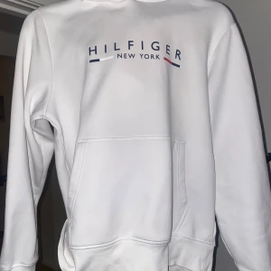 Vit hoodie från Hilfiger - Säljer en stilren vit hoodie från Hilfiger med texten 'Hilfiger New York' tryckt på bröstet i blått och rött. Hoodien har en klassisk design med känguruficka och långa ärmar. Perfekt för en avslappnad stil.