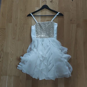 Vit klänning med paljetter - Säljer en söt vit klänning med glittrig paljettöverdel och volangkjol. Klänningen har smala axelband och en kort längd, perfekt för festliga tillfällen. Den är både elegant och lekfull med sina detaljer.