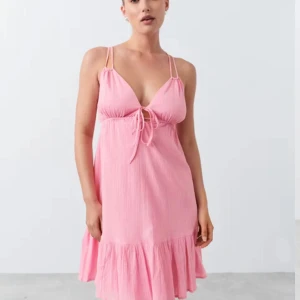 Rosa klänning med volangdetalj - Söt rosa klänning med smala axelband och v-ringning. Den har en knytning framtill och volangdetalj vid fållen. Perfekt för sommarens varma dagar. Helt slutsåld hos Gina tricot och har en fin öppen rygg men en X snörning 