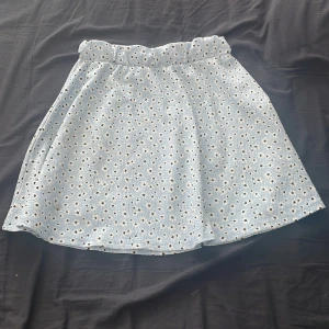 Ljusblå kjol med blommönster - Charmig ljusblå kjol med små blommor i vitt och svart. Kjolen har en elastisk midja för bekväm passform och är perfekt för varma dagar. Materialet är lätt och luftigt, vilket gör den idealisk för sommarens äventyr.