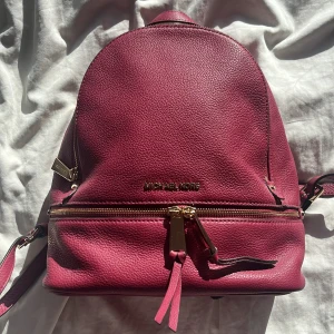 Vinröd ryggsäck från Michael Kors - Snygg vinröd ryggsäck från Michael Kors i skinn med guldfärgade dragkedjor. Perfekt för att bära dina dagliga nödvändigheter med stil. Rymligt huvudfack och en praktisk ytterficka med dragkedja. Justerbara axelremmar för bekväm passform.💞💞mått 31 x 27 x 13 cm. Väskan endast använd en gång.