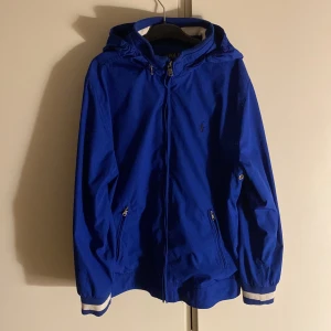 Ralph Lauren windbreaker - Snygg blå vindjacka från Ralph Lauren med huva och dragkedja. Jackan har långa ärmar med ribbade muddar och två fickor med dragkedja. Perfekt för blåsiga dagar! 9/10 skick!