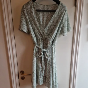 Grön blommig omlottklänning - Söt grön omlottklänning med blommigt mönster och knytband i midjan. Klänningen har korta ärmar och en v-ringad design som ger en avslappnad och somrig look.