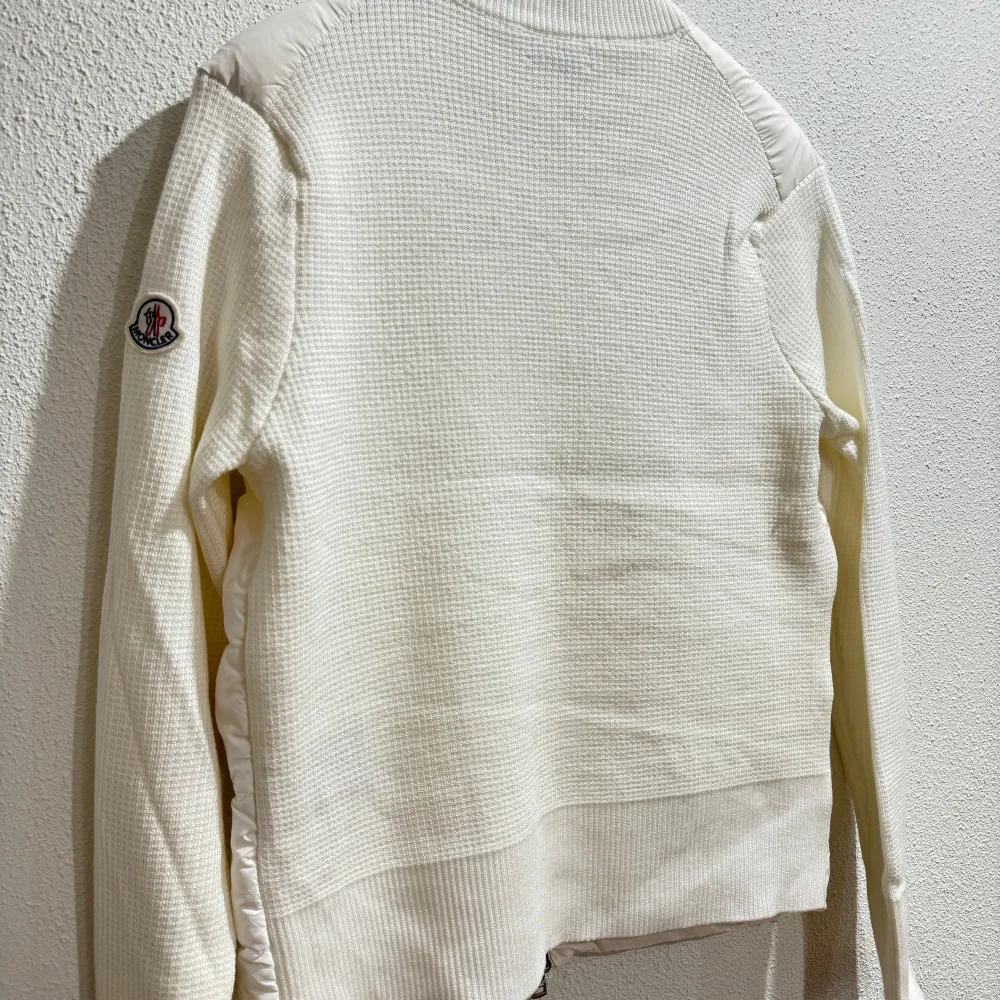 Säljer en stilren vit dunjacka från Moncler med dragkedja och broderad logotyp på ärmen. Jackan har en quiltad design framtill och är tillverkad i en kombination av syntet och stickat material. Perfekt för kyligare dagar med sin långa ärmar och bekväma passform. Taggar och lapp samt påse från Moncler medföljer. Takit.