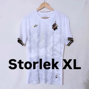Vit fotbollströja från Nike - Snygg vit fotbollströja från Nike med korta ärmar och stilrena detaljer i guld och svart. Tröjan har ett subtilt mönster och klubbmärke på bröstet. Perfekt för fotbollsfans som vill ha något unikt och stilrent.