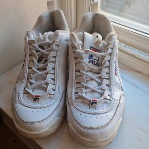 Vita sneakers från Fila - Säljer ett par vita Fila sneakers med snörning och märkets logotyp på sidan och baksidan. Skorna har en robust sula och är perfekta för en sportig look. De är tillverkade i syntetmaterial och har en klassisk design.