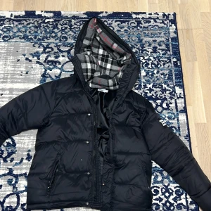 Sjuk Burberry - Säljer en snygg svart dunjacka från Burberry med en quiltad design och huva. Jackan har en dragkedja framtill och ett stilrent rutigt foder i huvan. Perfekt för kyliga dagar!