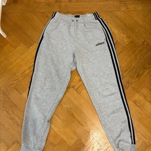 Grå mjukisbyxor från Adidas - Säljer ett par grå mjukisbyxor från Adidas med svarta ränder längs sidorna och elastisk midja. Byxorna har en avslappnad passform och är perfekta för en bekväm stil. Adidas-loggan är broderad på ena benet.