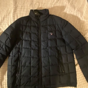 Svart quiltad jacka från GANT - Säljer en stilren svart quiltad jacka från GANT med broderad logga på bröstet. Jackan har dragkedja framtill och är perfekt för kyligare dagar. Den är långärmad och har en klassisk design som passar till det mesta. Den är en ganska ovanlig modell så den har ett värde, priset kan diskuteras 