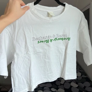 Vit croppad t-shirt från H&M - Säljer en vit croppad t-shirt från H&M med texten 'Recharge & Reset' i grönt och grått på framsidan. Perfekt för en avslappnad stil. T-shirten har korta ärmar, en rund halsringning och en oversized stil. Storlek: S. Axel: 50cm. Längd: 40cm. 💚
