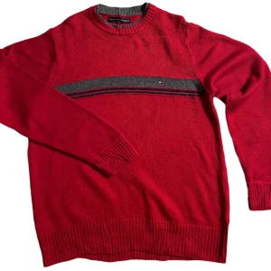 Röd tröja från Tommy Hilfiger - Snygg röd tröja från Tommy Hilfiger med grå detaljer runt halsen och ett diskret randigt mönster över bröstet. Tröjan har långa ärmar och ribbade muddar vid ärmslut och nederkant. Perfekt för en stilren look. Saknar storleks lapp