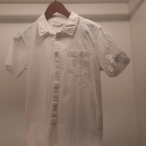 Vit kortärmad skjorta från 157 - Säljer en stilren vit kortärmad skjorta från 157. Skjortan har en klassisk krage och en bröstficka. Perfekt för en avslappnad och snygg look. Knapparna framtill ger en tidlös stil. Passar bra till både jeans och chinos.