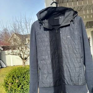 Cardigan från J.Lindeberg - Stilren  marin blå Cardigan Jacka från J.Lindeberg med quiltad framsida och dragkedja. Tidigare köpt från J Lindberg butiken i Täby centrum för 3599 och säljer för 999. Pris ej hugget i sten. Jackan har en huva med snörning och långa ärmar i ett stickat material. Perfekt för kommande vår dagar.