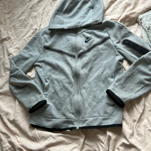 Grå Nike tech. - Säljer en grå huvtröja från Nike med dragkedja och svart logga på bröstet, den är i barnstorlek. Tröjan har en praktisk ficka med dragkedja på ärmen och svarta detaljer vid ärmslut och nederkant. 