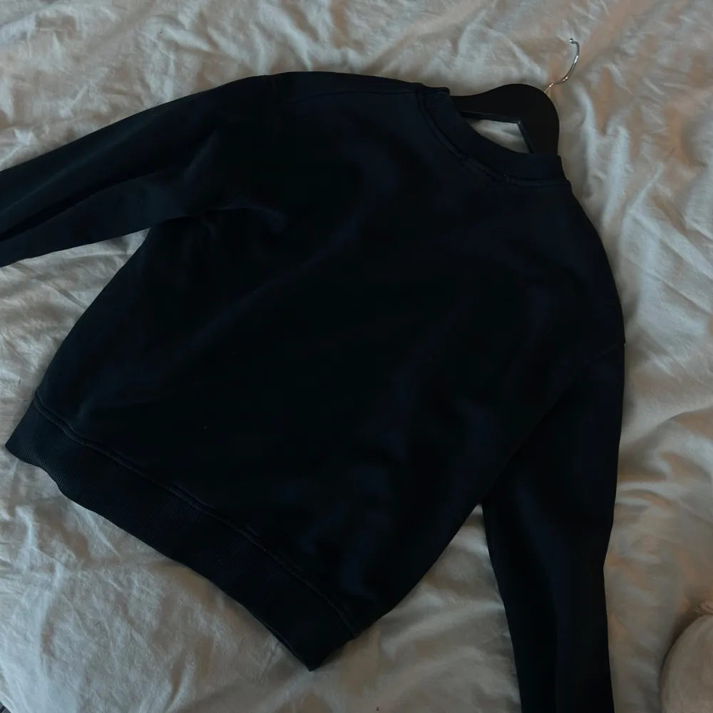 Säljer en stilren svart sweatshirt från NA-KD med broderad logga på bröstet. Tröjan har långa ärmar och ribbad krage samt muddar. Perfekt för en avslappnad look.. Neuletakit & Villapaidat.