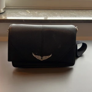 Svart axelväska från Zadig & Voltaire - Snygg svart axelväska från Zadig & Voltaire med ett coolt vingemblem på framsidan. Väskan har en justerbar axelrem och är tillverkad i skinn. Perfekt för att ge din outfit en edgy touch.
