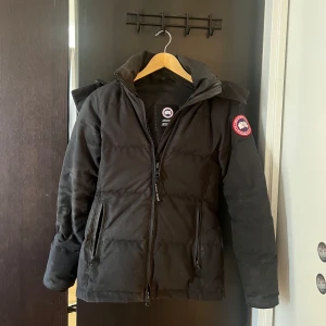 Svart Canada Goose dunjacka  - Säljer en svart dunjacka från Canada Goose med pälsdetalj på huvan. Jackan har en dragkedja framtill och en ikonisk logga på ärmen. Perfekt för kalla vinterdagar. Nypris 10000, säljer då den inte kommer till användning längre. Tyvärr har kvittot kommit bort pga flytt.