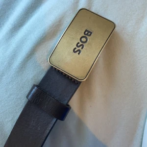 Svart bälte med guldfärgat spänne från Boss - Snyggt svart bälte i skinn med ett rektangulärt guldfärgat spänne från Boss. Perfekt för att ge en stilren touch till din outfit. Spännet har en elegant design med Boss-loggan ingraverad.