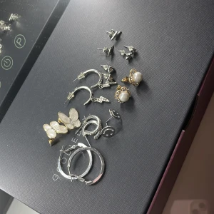 Set med örhängen i olika stilar - Ett set med olika örhängen, inklusive silverfärgade hoops, pärlörhängen med gulddetaljer, fjärilsformade örhängen och små stiftörhängen i olika former. Perfekt för att mixa och matcha till olika tillfällen.