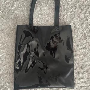 Svart glansig väska från Monki - Snygg svart glansig  väska från Monki. Perfekt för att bära med sig allt man behöver. Väskan har två handtag och en enkel design som passar till många olika stilar.