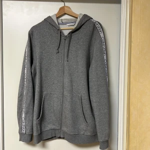 Grå hoodie från Calvin Klein - Säljer en stilren grå hoodie från Calvin Klein med dragkedja och huva. Tröjan har långa ärmar och märkets logga längs ärmarna. Perfekt för en avslappnad look.